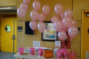 A hallway table is decorated with pink balloons and two buckets filled with paper “kindness fortunes” that children can draw from at the start of the school day.