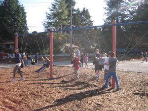 A group of elementary children swings together on a multi‑seat swing set under a bright, sunny sky, illustrating inclusive, peer‑led play.