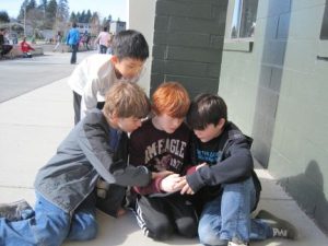 Four elementary school-aged boys crouched on the spavement outside a school, leaning over an iPad and listening intently to the audio being played