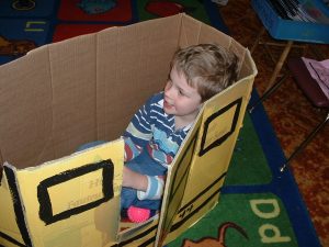 Ges sits happily inside a cardboard box painted like a train car, placed on the alphabet carpet in a kindergarten classroom.