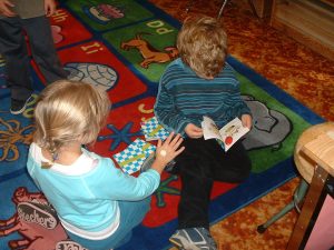A boy and a girl sit on a colorful alphabet carpet; the boy looks at a book while the girl uses a peer‑led strategy she learned to invite him to play.