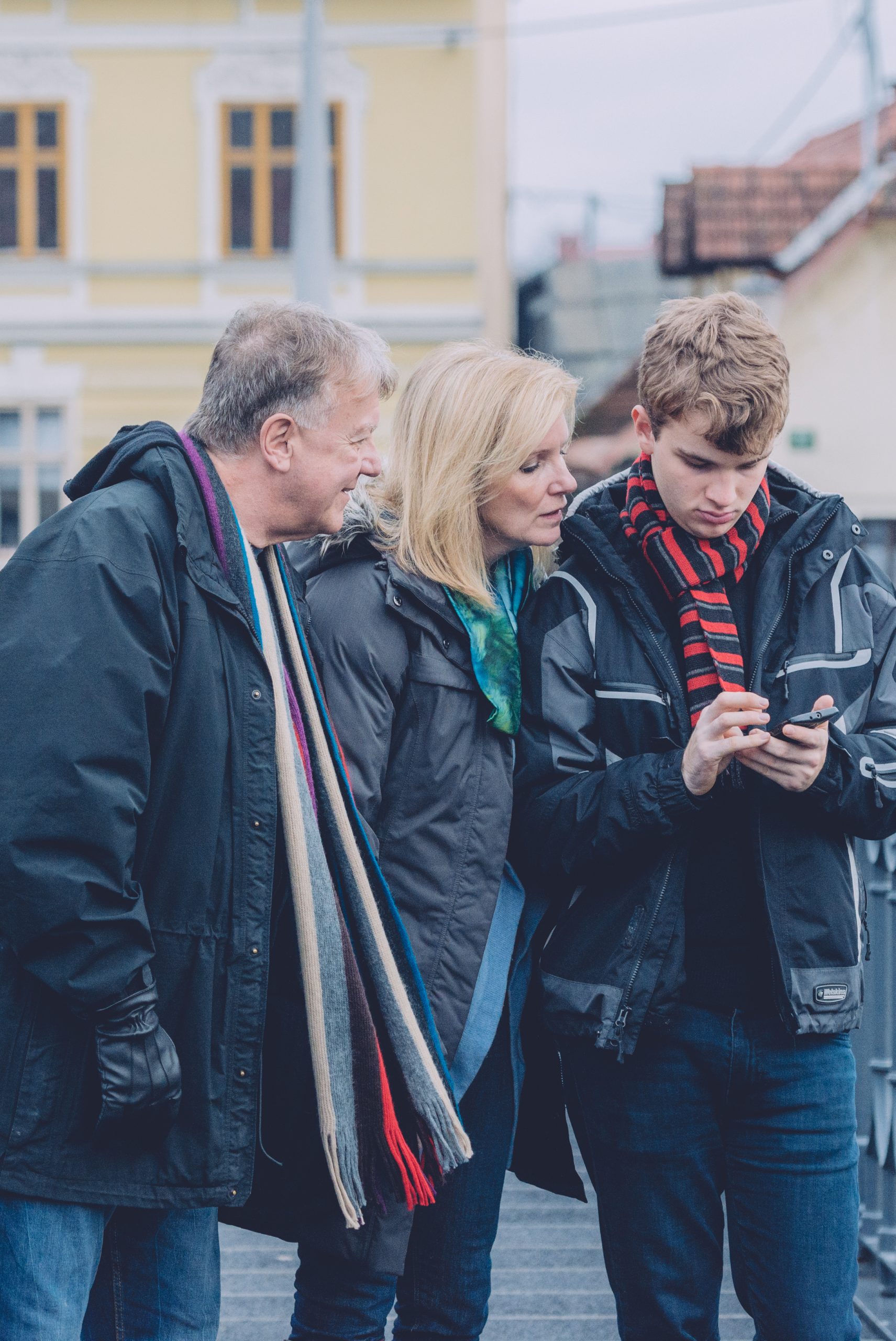 A young man using a speech‑assist app on his phone while an older man and woman watch attentively, illustrating public‑space communication and the social model of disability